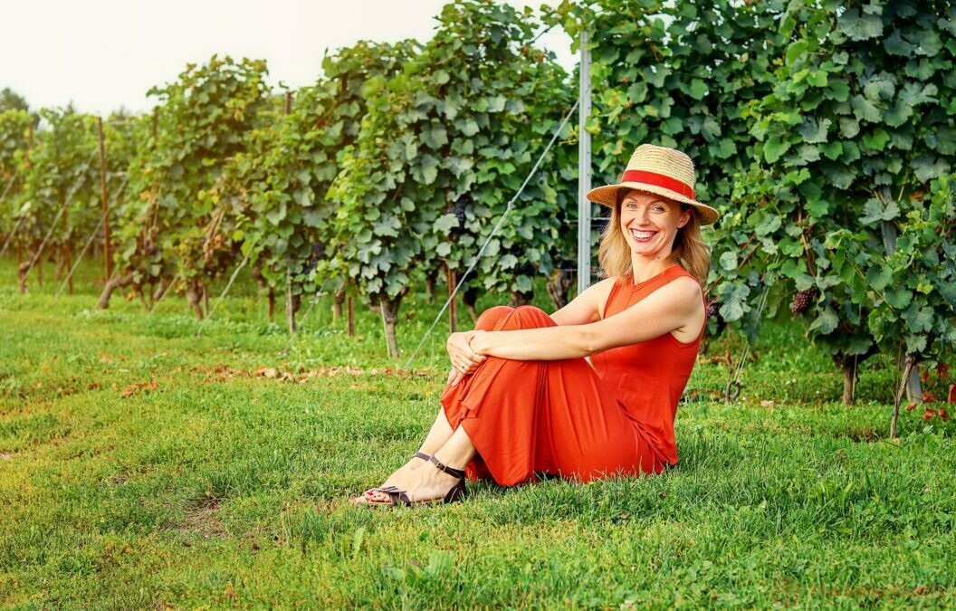 beautiful happy woman in red dress and straw hat sitting on green grass in vineyard smiling looking